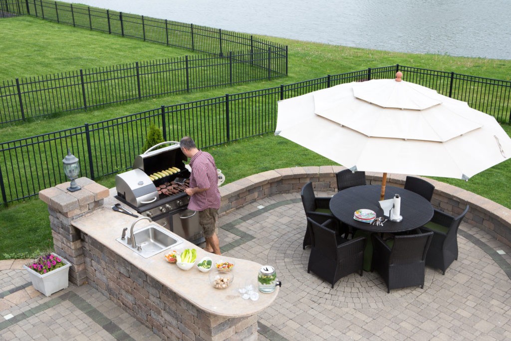28265194 - high angle view of a man cooking meat on a gas bbq standing in the sunshine on a paved outdoor patio at the summer kitchen preparing for guests with a table and chairs with a garden umbrella alongside